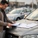 Un homme examine un carnet d’entretien vierge et des factures sans inscription posés sur le capot d’une voiture d’occasion, en lumière naturelle.