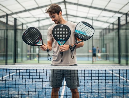 Un joueur amateur de padel compare trois raquettes de formes ronde, goutte d’eau et diamant sur un court couvert éclairé par la lumière naturelle.