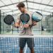 Un joueur amateur de padel compare trois raquettes de formes ronde, goutte d’eau et diamant sur un court couvert éclairé par la lumière naturelle.